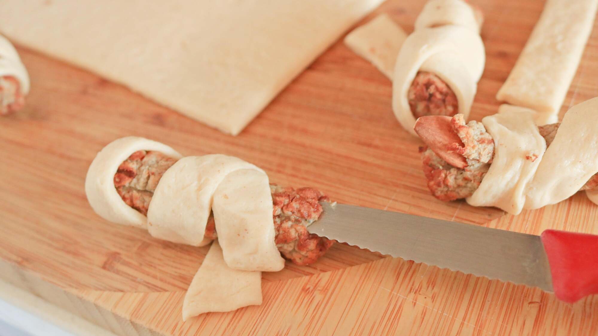 A paring knife cutting the end of a sausage wrapped in dough.